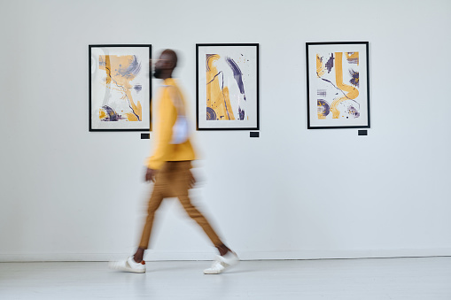 Blurred motion of African young man walking along art gallery with pictures on the wall