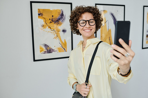 Happy young girl in eyeglasses using her smartphone for streaming while visiting art gallery