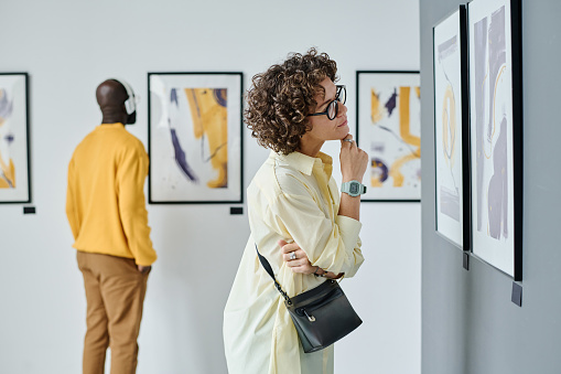 Young woman in eyeglasses examining modern art at gallery with other visitors