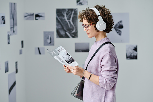 Young woman in wireless headphones reading booklet while standing at art gallery