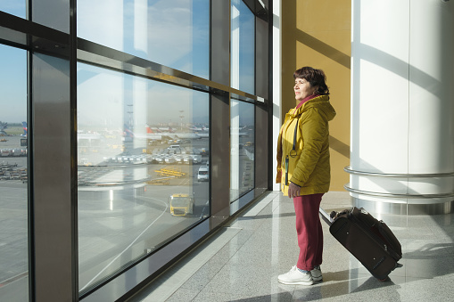 Full length portrait of an elderly woman with a suitcase. Mature brunette woman at the airport looks out the window at the runway while waiting for her flight.