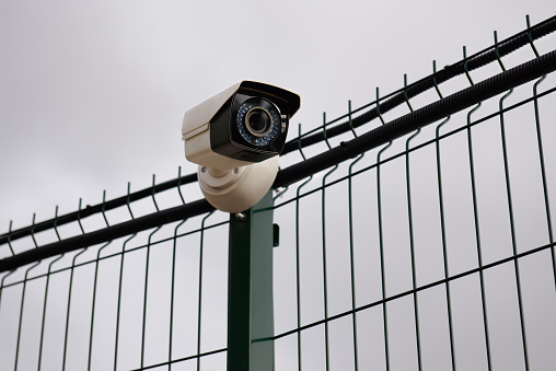 A CCTV surveillance camera on an iron fence against the background of clouds. Close-up.