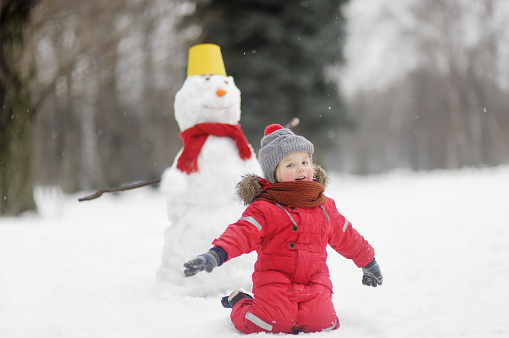 Little boy in red clothes having fun with big snowman. Child during stroll in a snowy winter park. Active winter outdoor leisure for family with children.