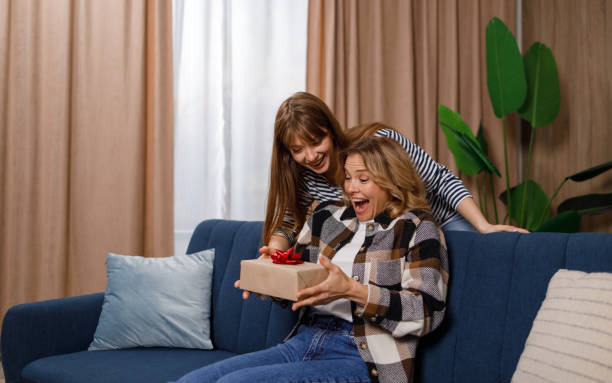 mama siedząca na kanapie w salonie jest zachwycona prezentem córki. dorosła dziewczyna gratuluje mamie wakacji - birthday present family facial expression birthday zdjęcia i obrazy z banku zdjęć