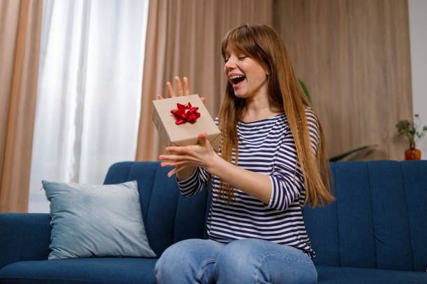 istock Joyful young woman holding a gift box with present while sitting on sofa in living room 1432877518