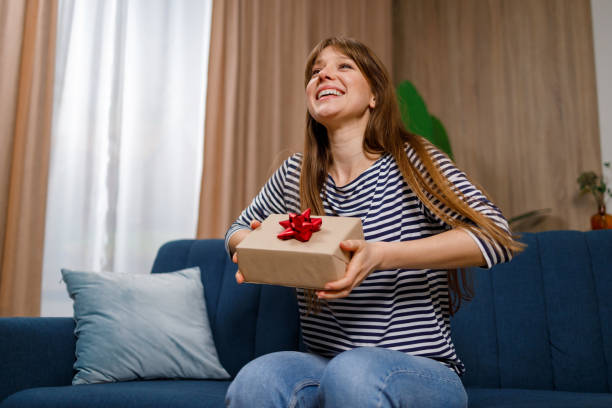 istock Delighted young woman holding a gift box with present while sitting on sofa in living room 1432877519