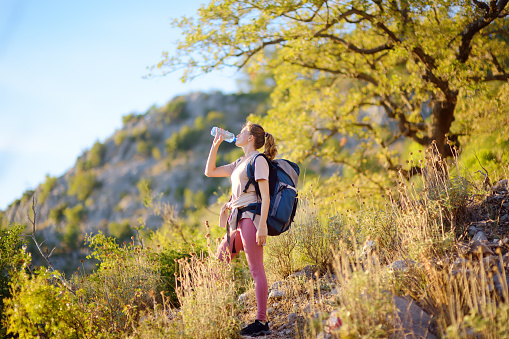 Young woman hiking in mountains in Europe at summer. Girl drinking water on hot day. Sunset landscape of hills. Concepts of adventure, extreme survival, orienteering. Solo journey. Backpacking hike
