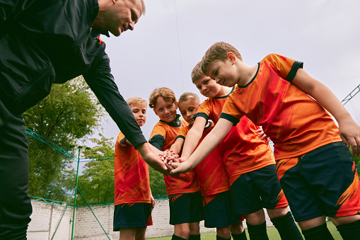 Determined to win. Junior soccer team stacking hands before a match, outdoors. Little boys, athletes in black-orange football kits. Concept of sport, competition, teamwork and achievements