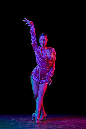 Queen. Portrait of young graceful flexible woman dancing ballroom dance without partner isolated on dark background in neon light. Concept of art, sport, beauty, music, style