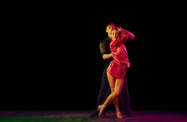 Photo of Full-length portrait of young beautiful man and woman dancing ballroom dance isolated over dark background in neon light. Concept of art, beauty, grace, action, emotions.