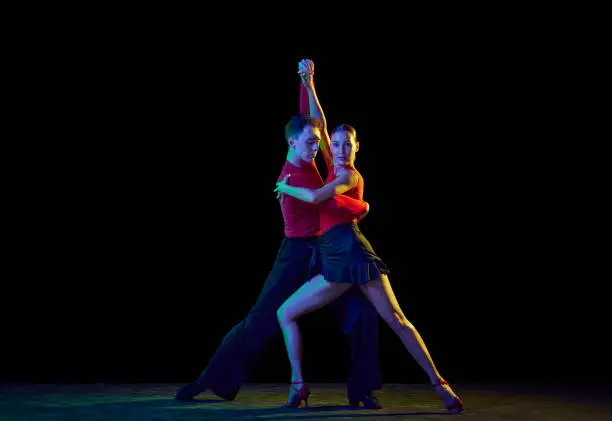 Photo of Full-length portrait of young beautiful man and woman dancing ballroom dance isolated over dark background in neon light. Concept of art, beauty, grace, action, emotions.