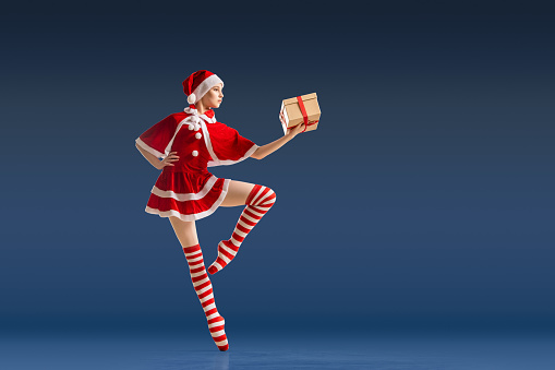 Dancing ballerina with gift in her hands on pointe shoes in costume of Santa Claus on dark blue background.