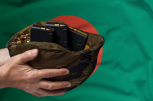 A military helmet with cartridges and magazines for a rifle in the hands of a man against the background of the flag of Bangladesh. The concept of selling weapons or military assistance.
