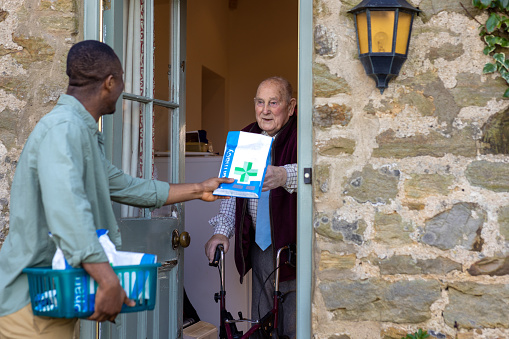 Three-quarter-length shot of a senior man standing at his front door to a pharmacist who is delivering a prescription wearing casual clothing. The man accepts the package smiling back at the delivery person. They are in the Northeast of England.