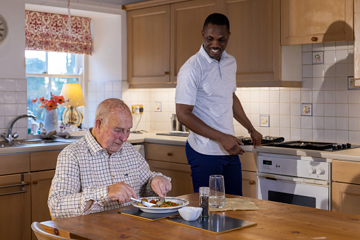 Medium shot of a senior man being eating his dinner that has been served by a male healthcare worker.  The healthcare worker is smiling while he looks over at the senior man. They are in the Northeast of England.