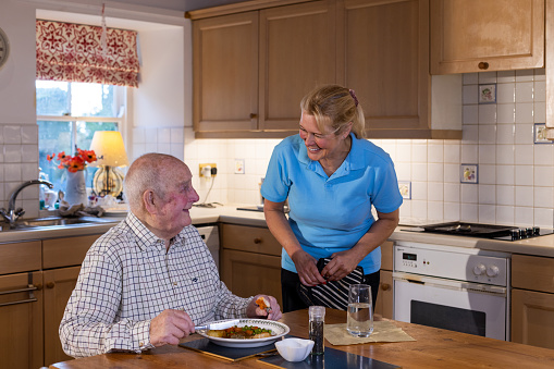 Medium shot of a senior man sitting and having his dinner served to him by his carer who is talking to him in the kitchen in the Northeast of England.