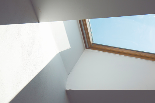 Dormer wooden window in the white sloping ceiling. Square ceiling window. Looking up to the blue sky