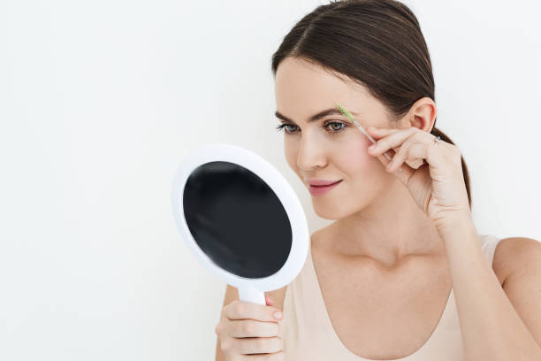 istock Beautiful woman combing her eyebrows with brush tool while looking into portable mirror, isolated on white. Eyebrow brushing process 1432899535