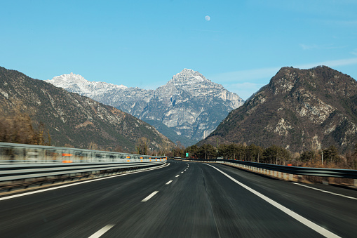 driving on the highway in a valley between the mountains, with clear sky and the visible moon, in the afternoon during winter season. highway section between Carnia and Chiusaforte, district of Udine, FVG region, Italy