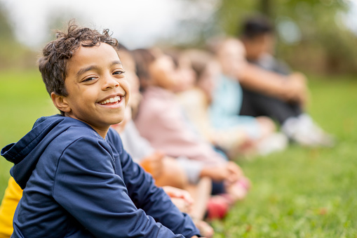 A young mixed race boy sits in the grass with his peers as they take a break after a Cross Country Race.  The children are each sitting side-by-side with their legs crossed and the young boy is looking to the camera with a smile.