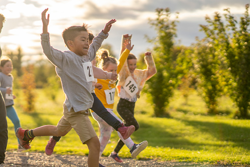 A small group of children raise their hands in the air as they cross the finish line first during a Cross Country race. They are each dressed comfortably in athletic wear and are smiling as they proudly cross the finish line.