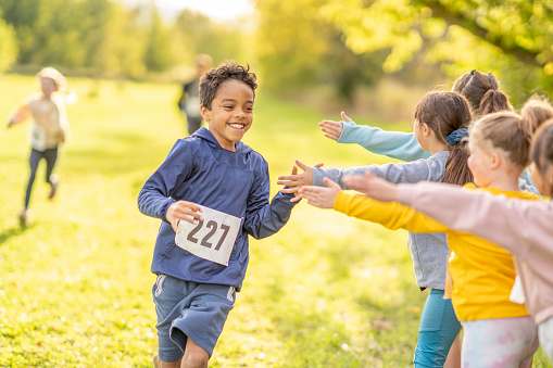 A young boy holds out his hand to high-five his peers on the sidelines as he finishes a Cross Country Race strong.  He is dressed comfortably and has a numbered bib on as he smiles and finishes the race.