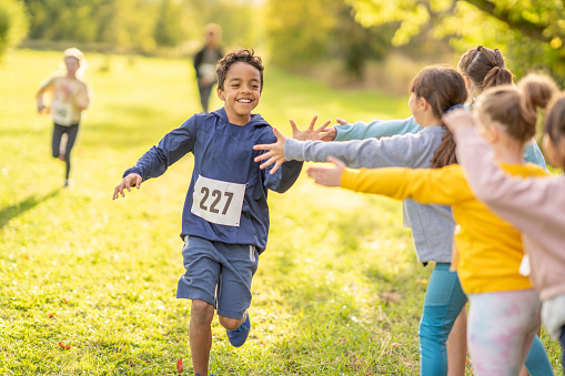A young boy holds out his hand to high-five his peers on the sidelines as he finishes a Cross Country Race strong.  He is dressed comfortably and has a numbered bib on as he smiles and finishes the race.