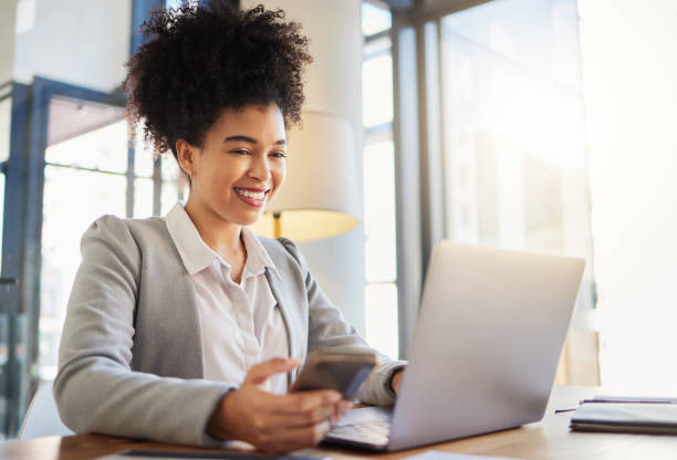 istock Happy corporate employee texting on a phone while working on a laptop in an office, reading email. Young professional assistant multitasking to complete an online task, efficient time management 1432907531