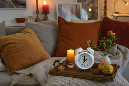 White vintage alarm clock showing fall back hour in autumn after daylight saving time, wooden tray with candle and warm colored fall decoration on a cozy couch, copy space, selected focus