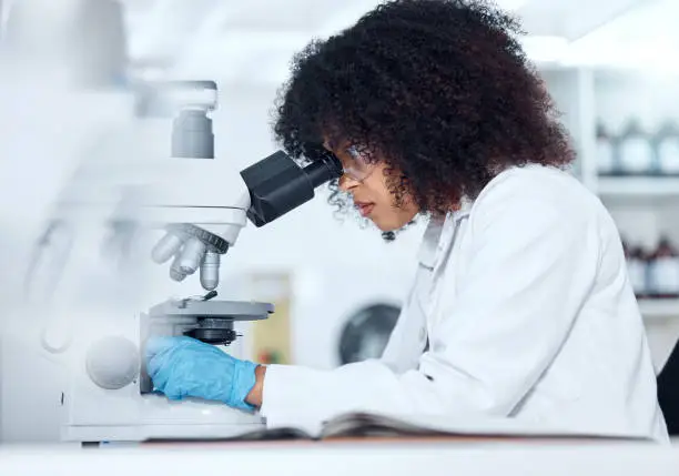 Photo of One mixed race scientist with curly hair wearing safety goggles and gloves analysing medical test samples on a microscope in a lab. Young woman doing forensic research and experiment to develop a cure