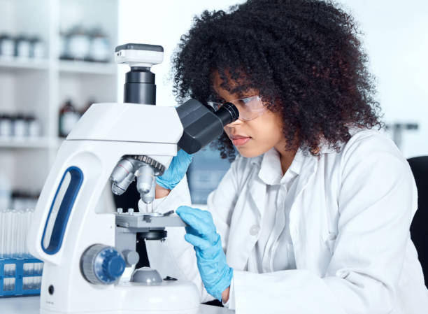 junge afroamerikanerin in einem laborkittel und einer schutzbrille, die medizinische proben auf einem mikroskop in ihrem labor betrachtet. eine gemischtrassige wissenschaftlerin mit brillen und handschuhen, die forschung betreibt - laboratory microscope pathologist scientist stock-fotos und bilder
