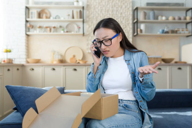 unzufriedene frau erhielt ein kaputtes paket, das von einem online-online-shop geliefert wurde, asiaten streiten sich mit dem kundendienst, sprechen mit dem betreiber am telefon, sitzen zu hause in der küche - ankunft stock-fotos und bilder