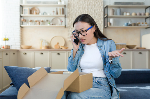 Unsatisfied woman received a broken parcel delivered from an online online store, Asian quarrels with the customer support service, talks to the operator on the phone, sitting home in the kitchen.