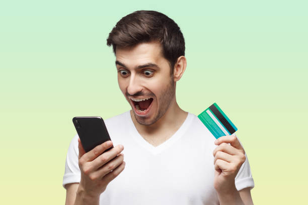 istock Excited handsome man with mouth open, holding credit card, using mobile phone on green background 1432926385
