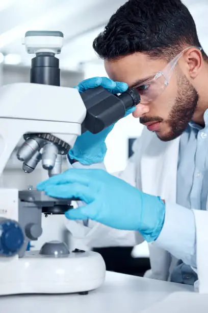 Photo of One mixed race scientist wearing safety goggles and gloves analysing medical test samples with a microscope in a lab. Young indian man developing a cure while doing forensic research and experiments