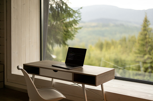 Workplace with laptop on table in front of panoramic window with scenic view on mountains and pine forest. Home office and remote work concept