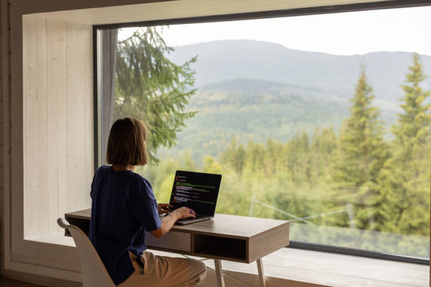 Woman works on laptop remotely in house on nature Woman works on laptop while sitting by the table in front of panoramic window with great view on mountains. Remote work and escaping to nature concept home office stock pictures, royalty-free photos & images