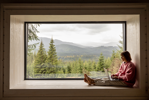 Woman works on laptop while sitting on a window with great view on mountains. Remote work from cozy place and escaping to nature concept