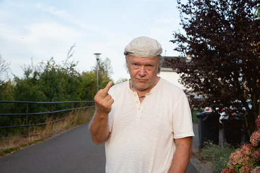 Senior man, wearing white t-shirt and cap, stannding outdoors on street, showing the finger
