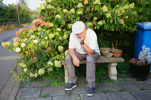 Senior man, wearing white t-shirt and cap, sitting on bench, head supprted by hand, seems to be depressed or exhausted