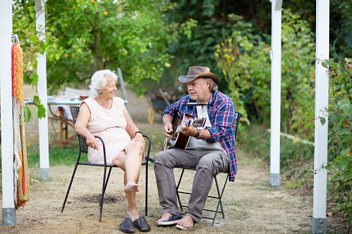 Senior man, wearing a leather hat and chequered shirt, sitting outdoors in yard and playing guitar, his wife, wearing a light pink dress, is sitting besides him, listening and smiling, the grass has dried out after months of heat without rain
