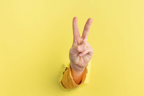 dziecięca ręka licząca i pokazująca dwa palce w górę przez otwór w żółtym papierze z podartymi krawędziami. gest pokoju lub zwycięstwo, znak v - peace sign counting child human finger zdjęcia i obrazy z banku zdjęć