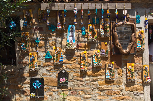 Gökçeada (Imbros), Turkey - August 3, 2022: Handmade decorative objects hanging on the old stone wall of a traditional Aegean house. Evil eye beads that protect from negative energy, art and craft works describing island life, authentic mirror. Souvenirs from Gökçeada (imbros). Horizontal close-up.