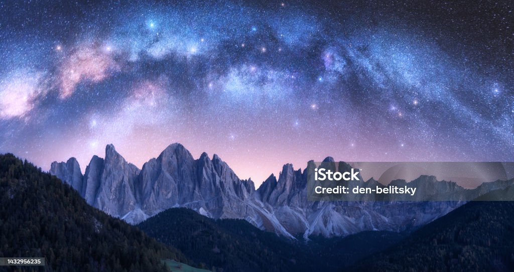 Acrhed Milky Way over beautifull rocks at starry night in summer in Dolomites, Italy. Purple sky with stars and bright milky way arch over high alpine rocky mountains. Space background. Nature Mountain Stock Photo