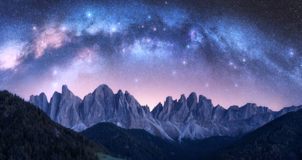 Acrhed Milky Way over beautifull rocks at starry night in summer in Dolomites, Italy. Purple sky with stars and bright milky way arch over high alpine rocky mountains. Space background. Nature stock photo