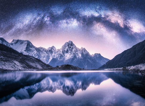 Milky Way arch over snowy mountains and lake at night. Landscape with snow covered high rocks, violet starry sky, reflection in water in Nepal. Sky with stars. Bright milky way in Himalayas. Space