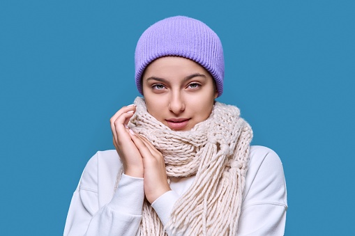 Winter headshot portrait of young female in hat sweater scarf. Adorable beautiful cute teenage girl looking at camera on blue studio background. Cold season New Year Christmas holidays fashion beauty