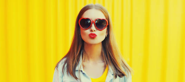 Summer portrait of beautiful young woman blowing her red lips sending sweet air kiss wearing heart shaped sunglasses on colorful yellow background stock photo
