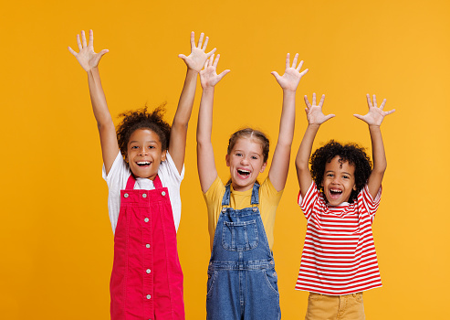 Group of cheerful happy multinational children shout with joy raising his hands up on colored yellow background