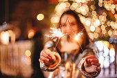 Young happy woman holding sparklers while standing against illuminated Christmas tree outdoors in city